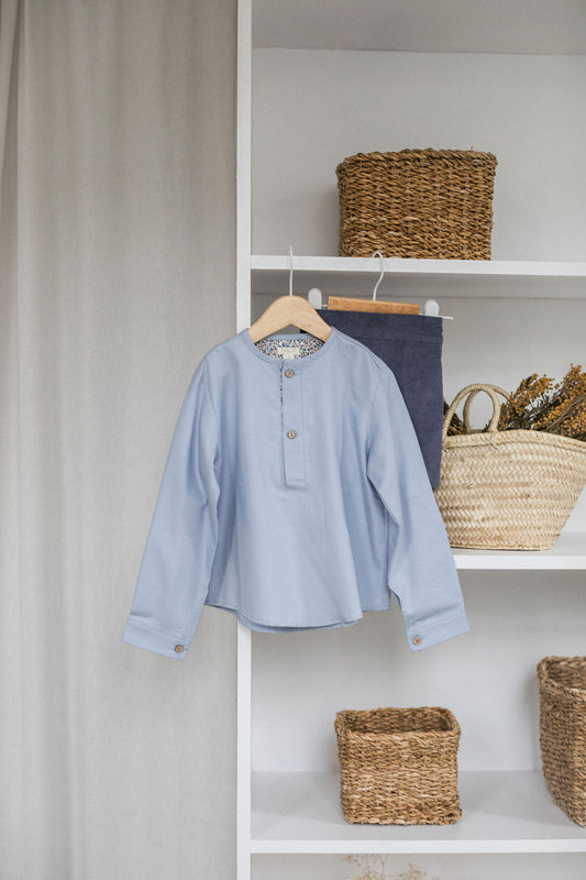 An arctic blue shirt with a mao collar and long sleeves, displayed on a hanger against a backdrop of wicker baskets and home decor items.
