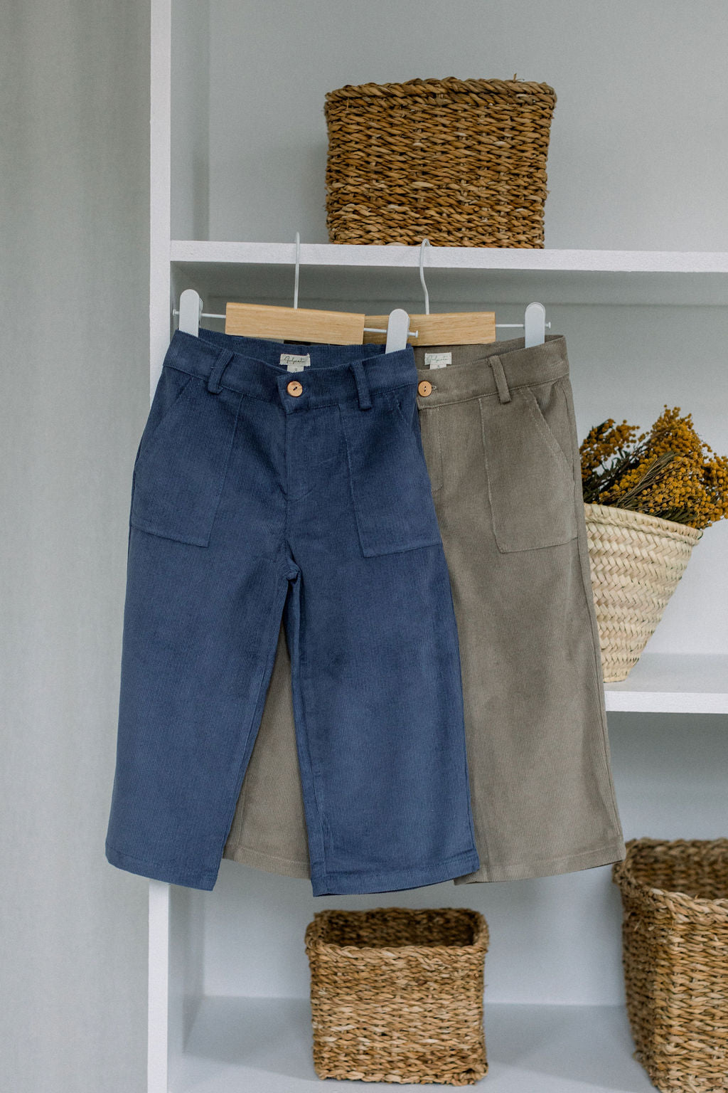 A pair of corduroy wide trousers displayed on a white hanger, with wooden button detailing, in a coconut color, alongside a woven basket and decorative branches.