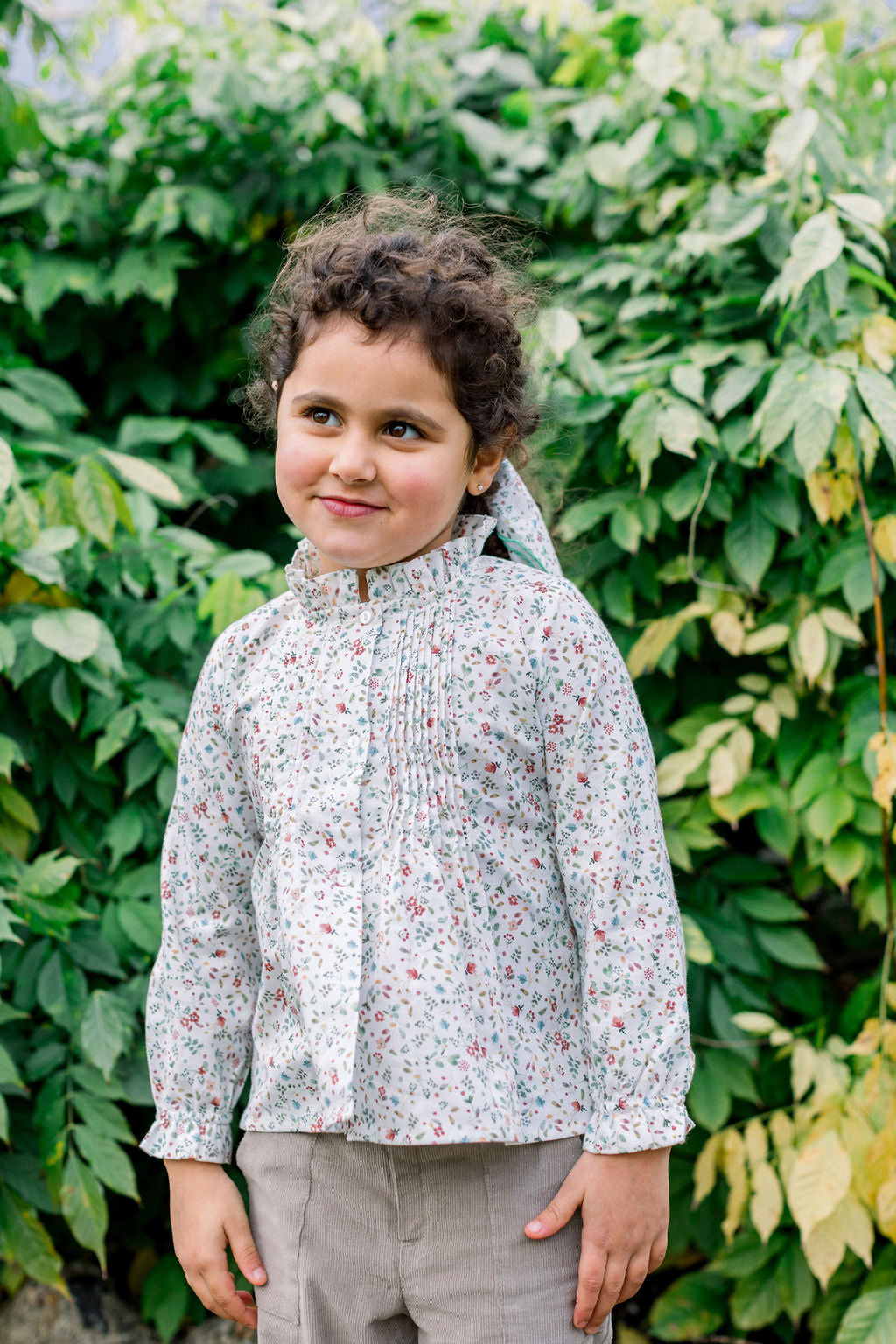 A child wearing a floral and leaves patterned blouse with a sweet ruffle collar and long sleeves with frill details, standing in front of a green leafy background.