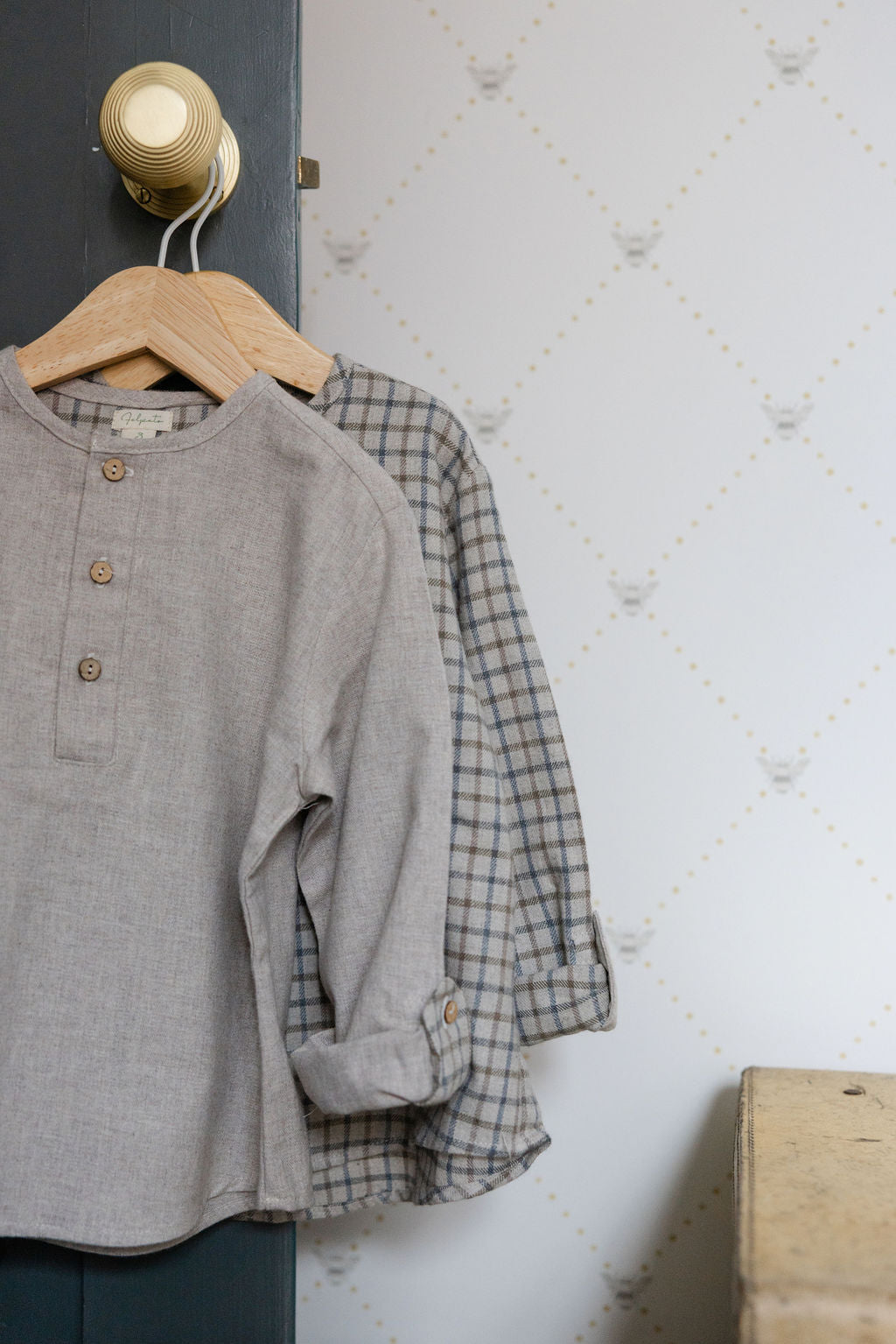 Gray shirt and plaid shirt hanging on a wooden hanger against a white wall.