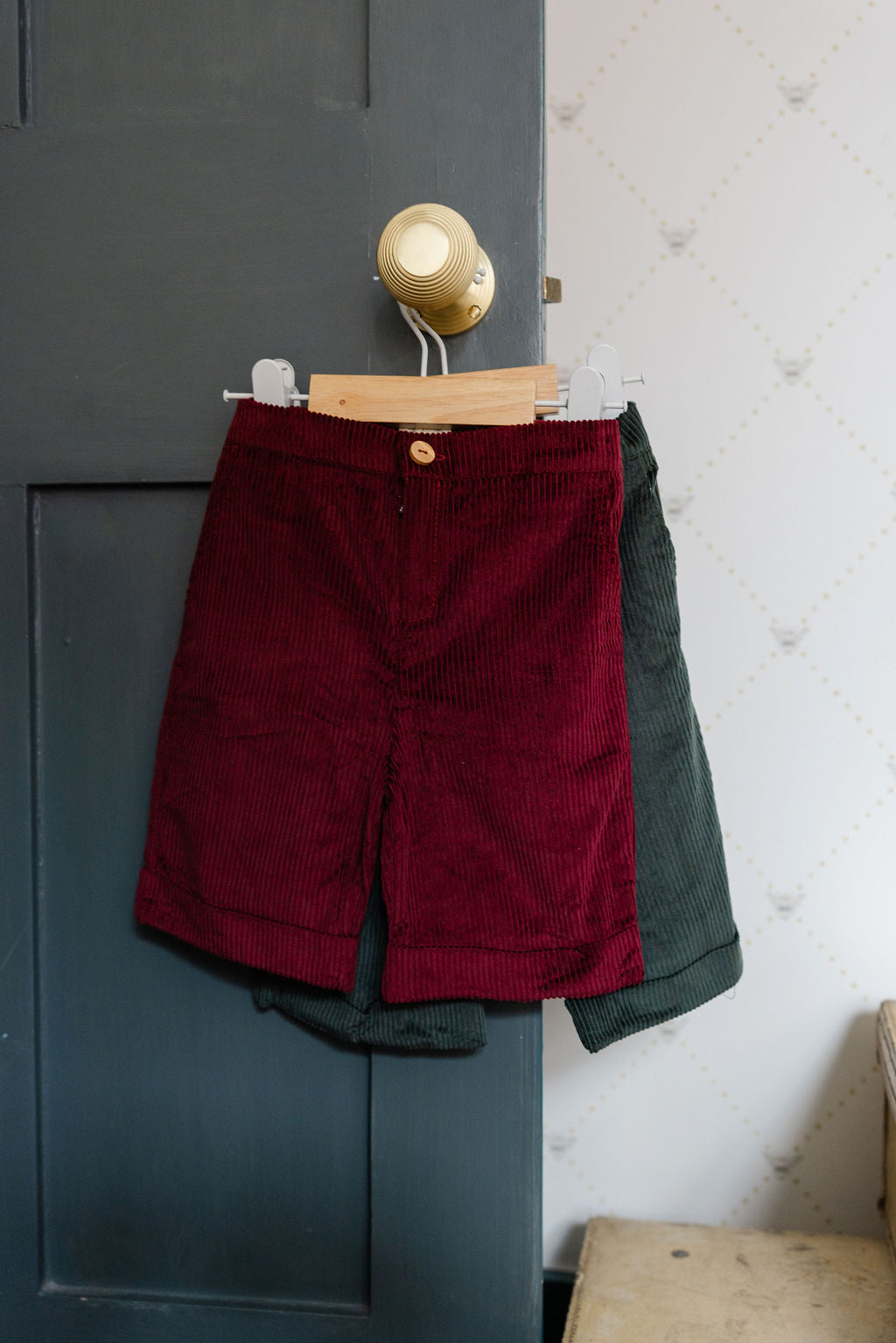 Red shorts and green jacket hanging on a hook against a gray wall.