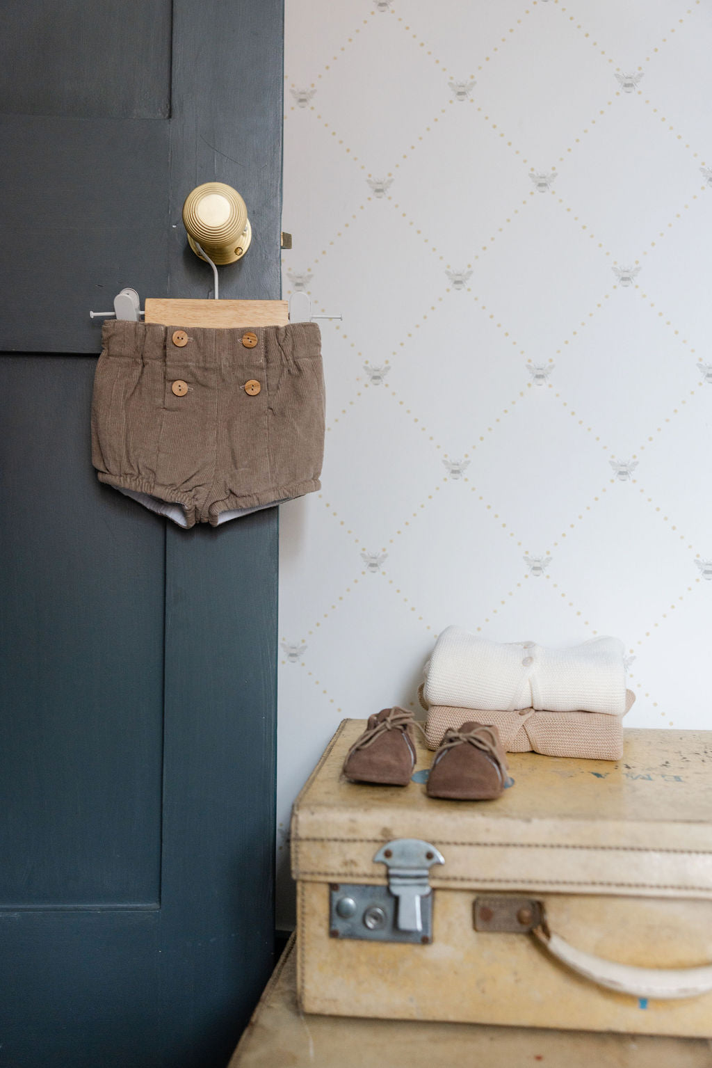 Brown shorts hanging on a door handle next to a wooden suitcase with shoes and clothes on a table.