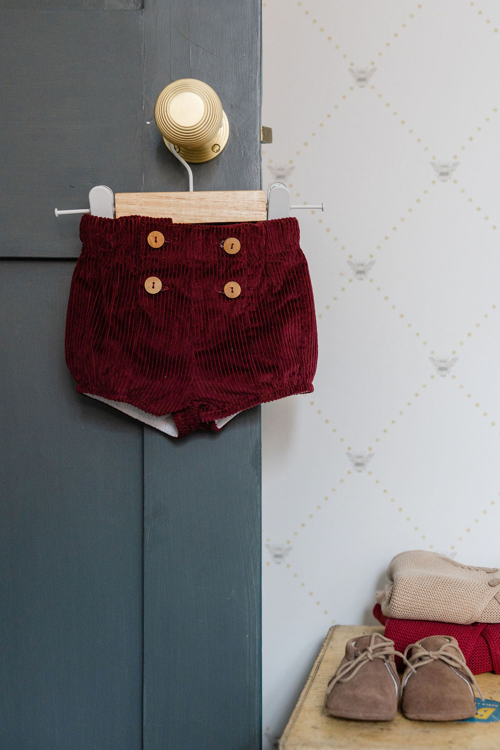 Red corduroy shorts with gold buttons hanging on a door next to a wall with a light fixture.
