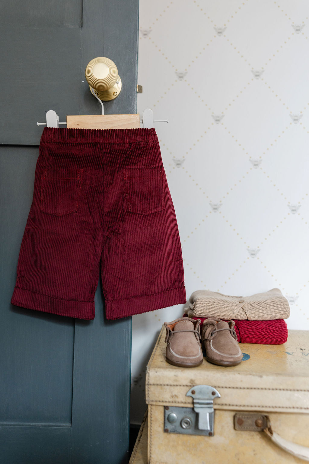 Red corduroy pants hanging on a hook with a suitcase and shoes below.