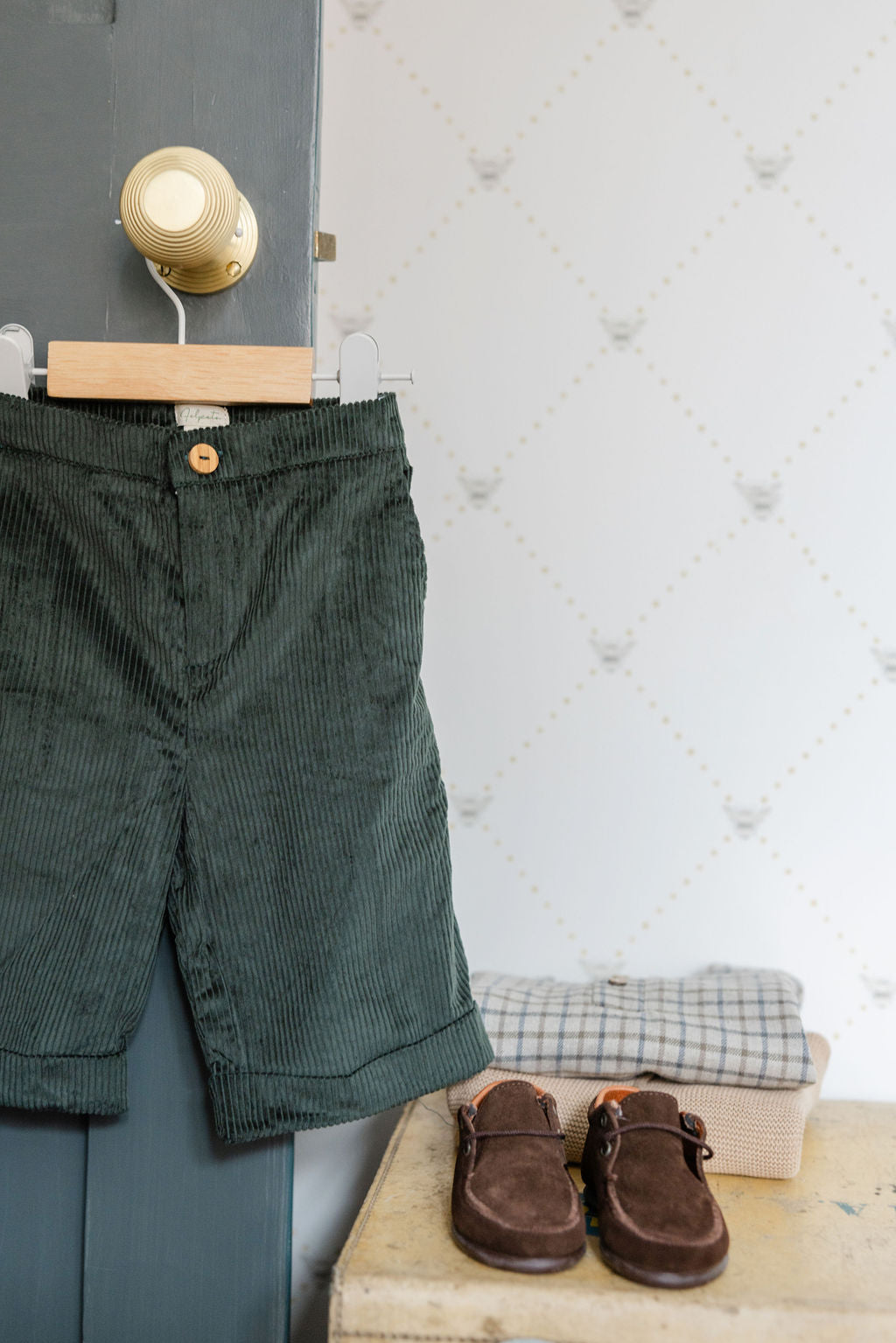 Green corduroy pants and brown shoes on a wooden surface with a white textured wall background.