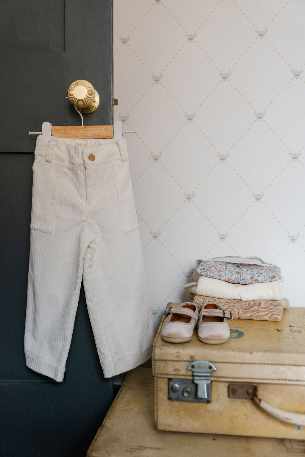 White pants hanging on a hook next to a stack of folded clothes and a vintage suitcase against a white wall.