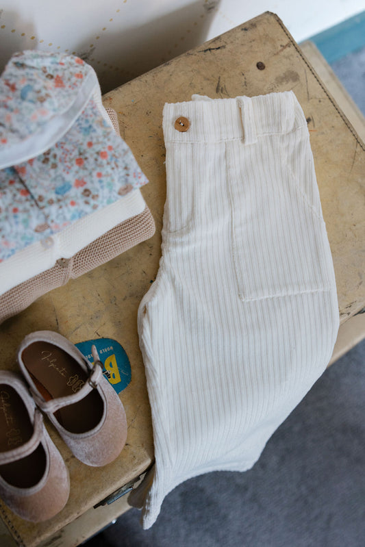 White ribbed pants with a button on a cardboard box with shoes and fabric in the background.