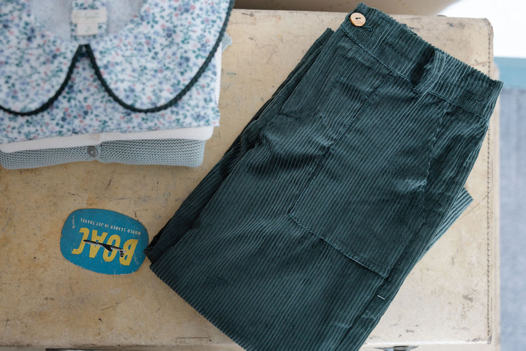 Green corduroy pants on a wooden surface with a floral-patterned garment in the background.