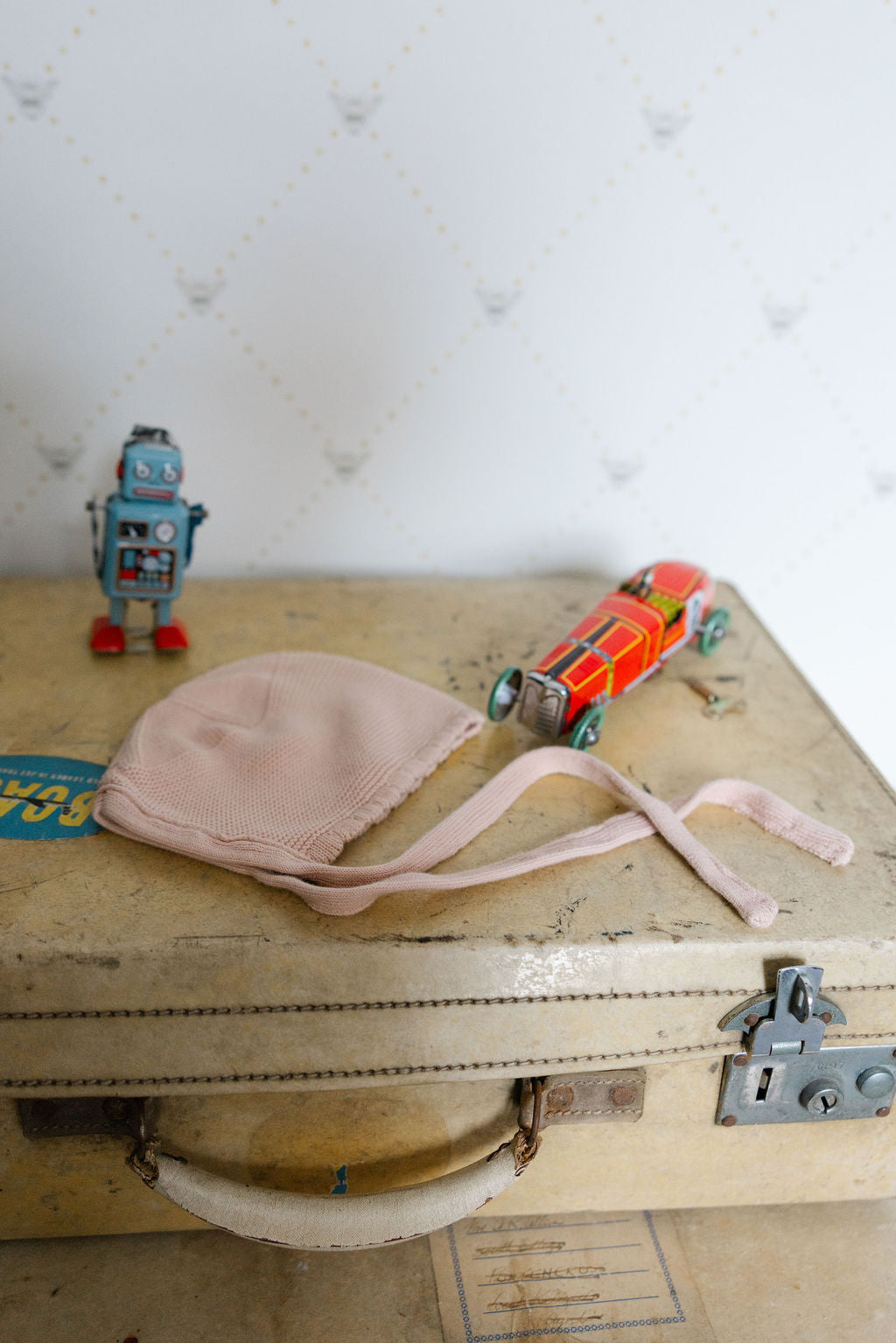 Vintage suitcase with a pink Bonet, red and orange toy car, and blue robot on a textured white background