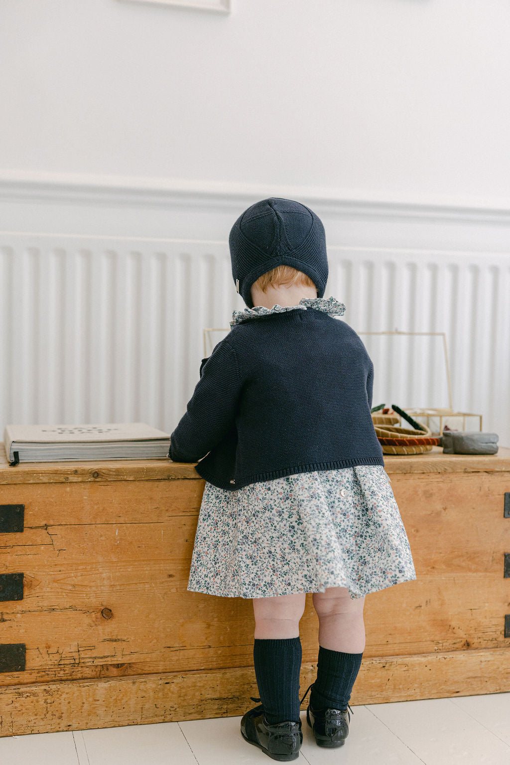Child wearing a navy blue coat and floral dress standing in front of a wooden dresser.