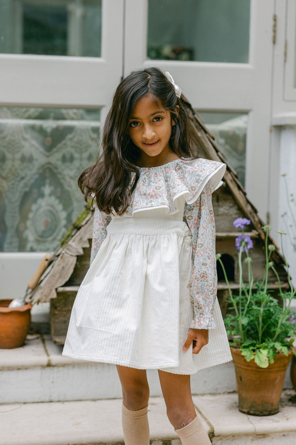 Young girl in a white dress standing outdoors with plants and a wooden structure in the background