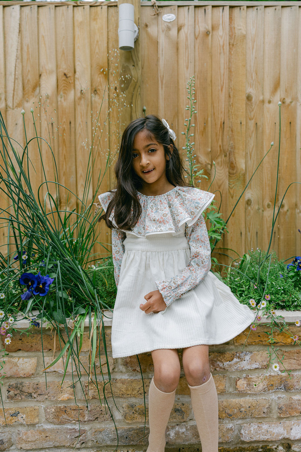 Young girl in a white dress standing in a garden with a wooden fence and plants in the background.