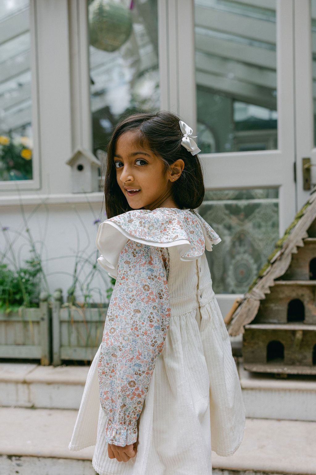 Young girl in a floral outfit standing outdoors with a greenhouse in the background