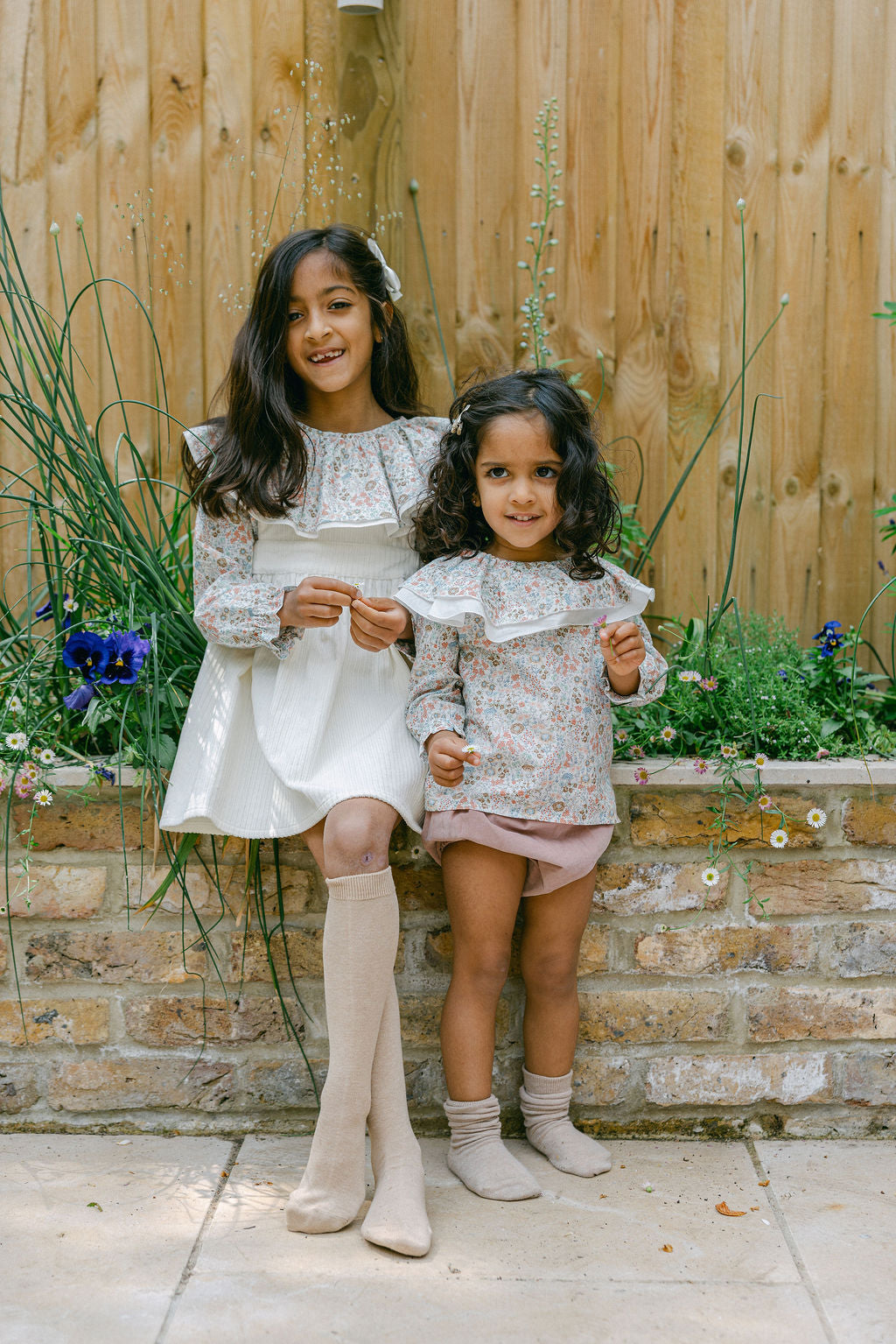 Two young girls in matching outfits standing outdoors against a wooden fence and stone wall.