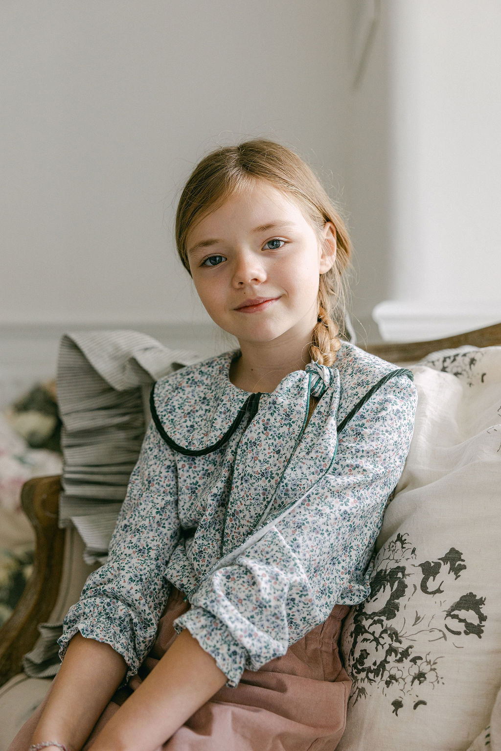 Young girl sitting on a couch wearing a floral blouse and pink skirt.