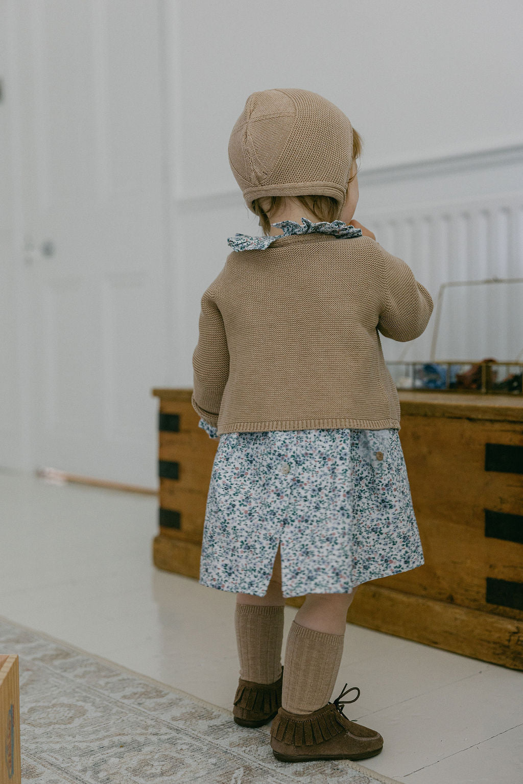Child wearing a brown coat and hat, standing in a room with wooden furniture.