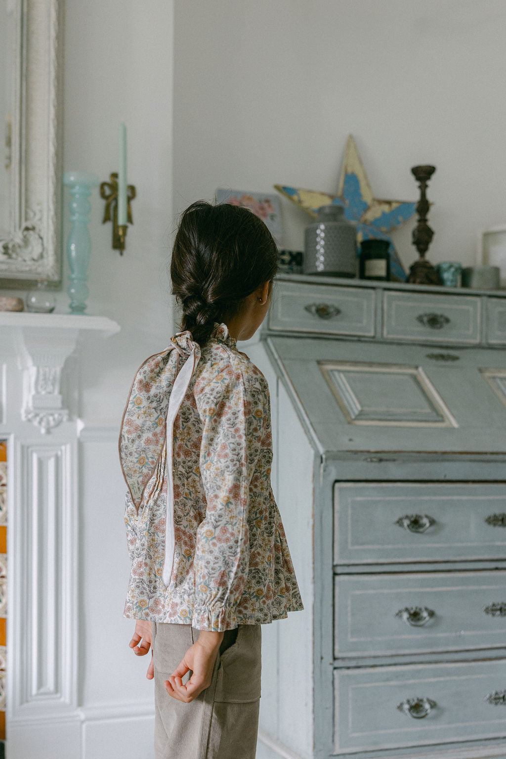 Child in a floral blouse standing in a room with a dresser and decorative items.