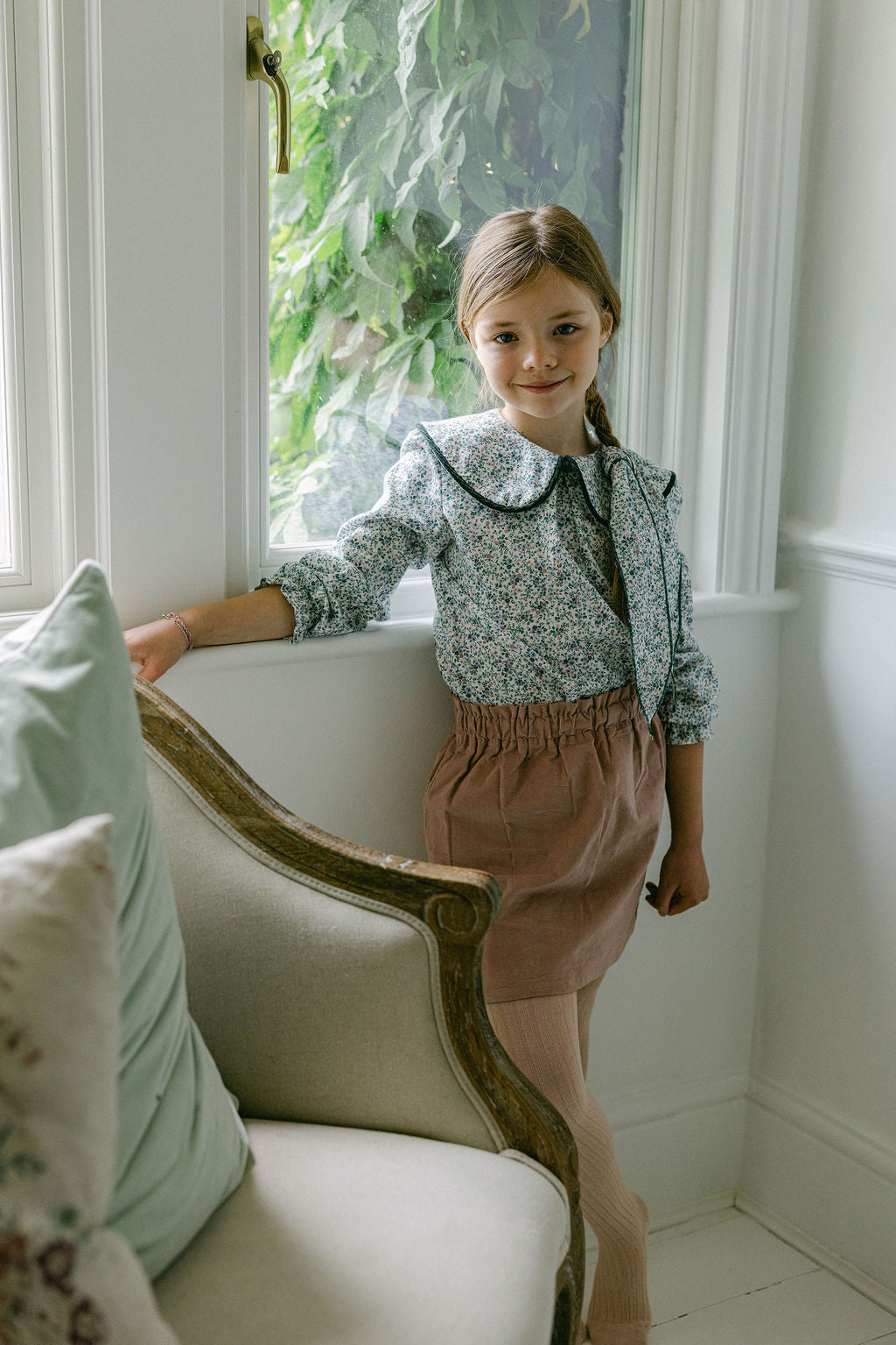Young girl standing by a window in a room with white walls and decorative elements.