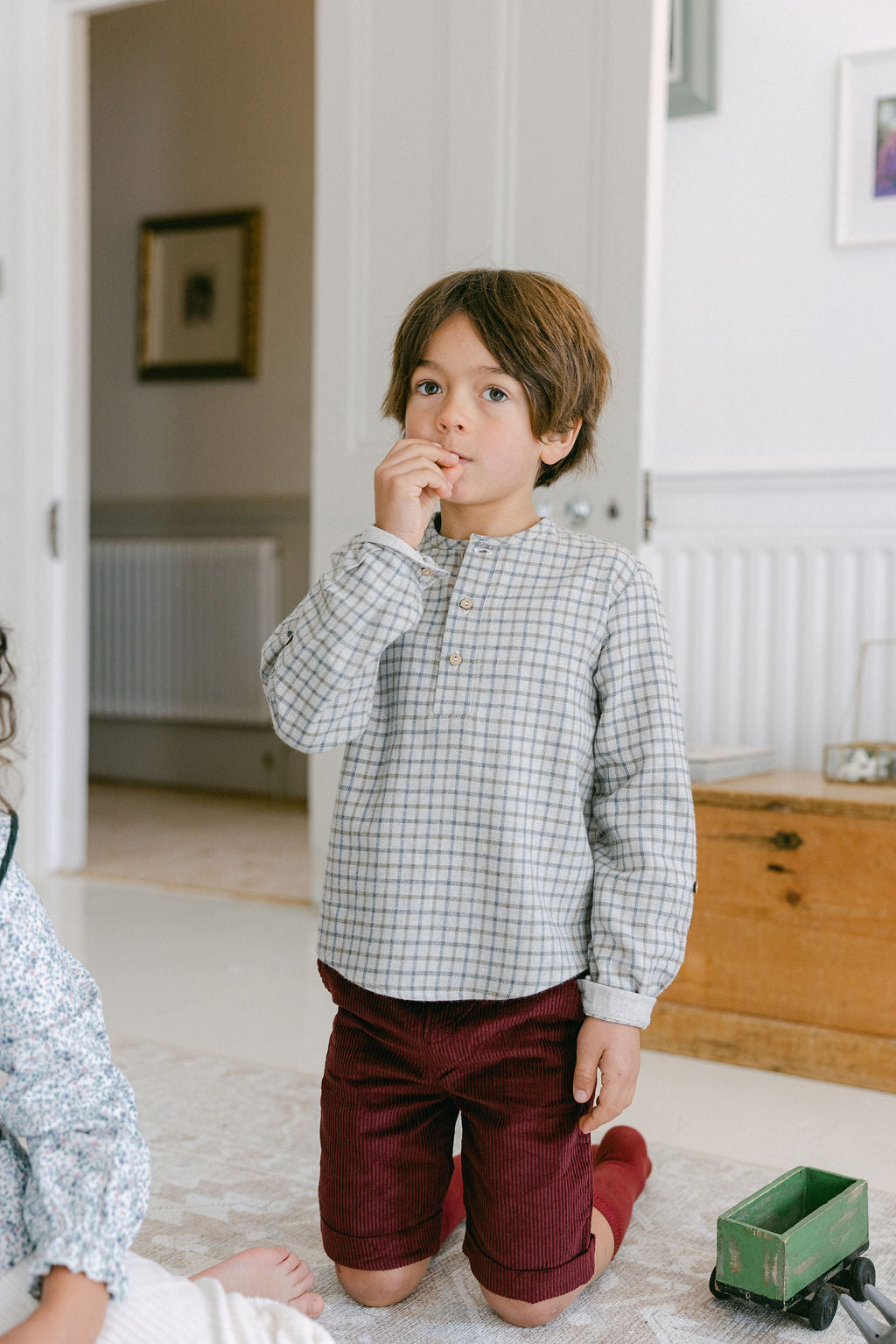 Child wearing a checkered shirt and red pants standing in a room with white walls and wooden furniture.