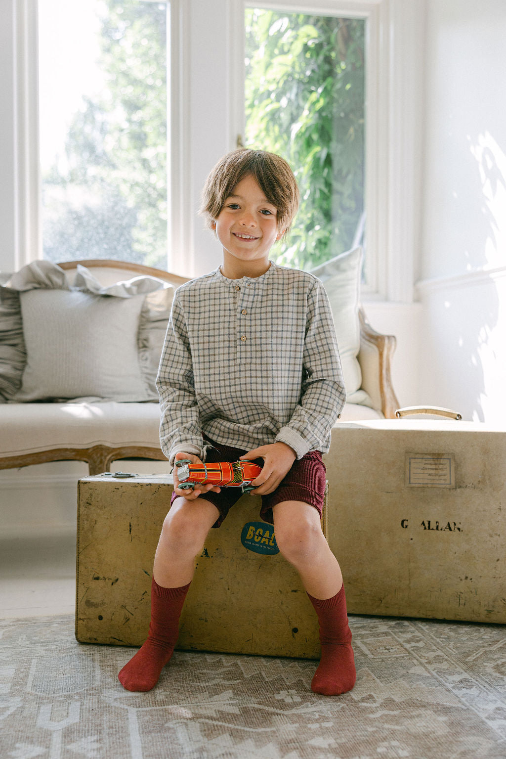 Child sitting on a vintage suitcase holding a toy train in a bright room with large windows.