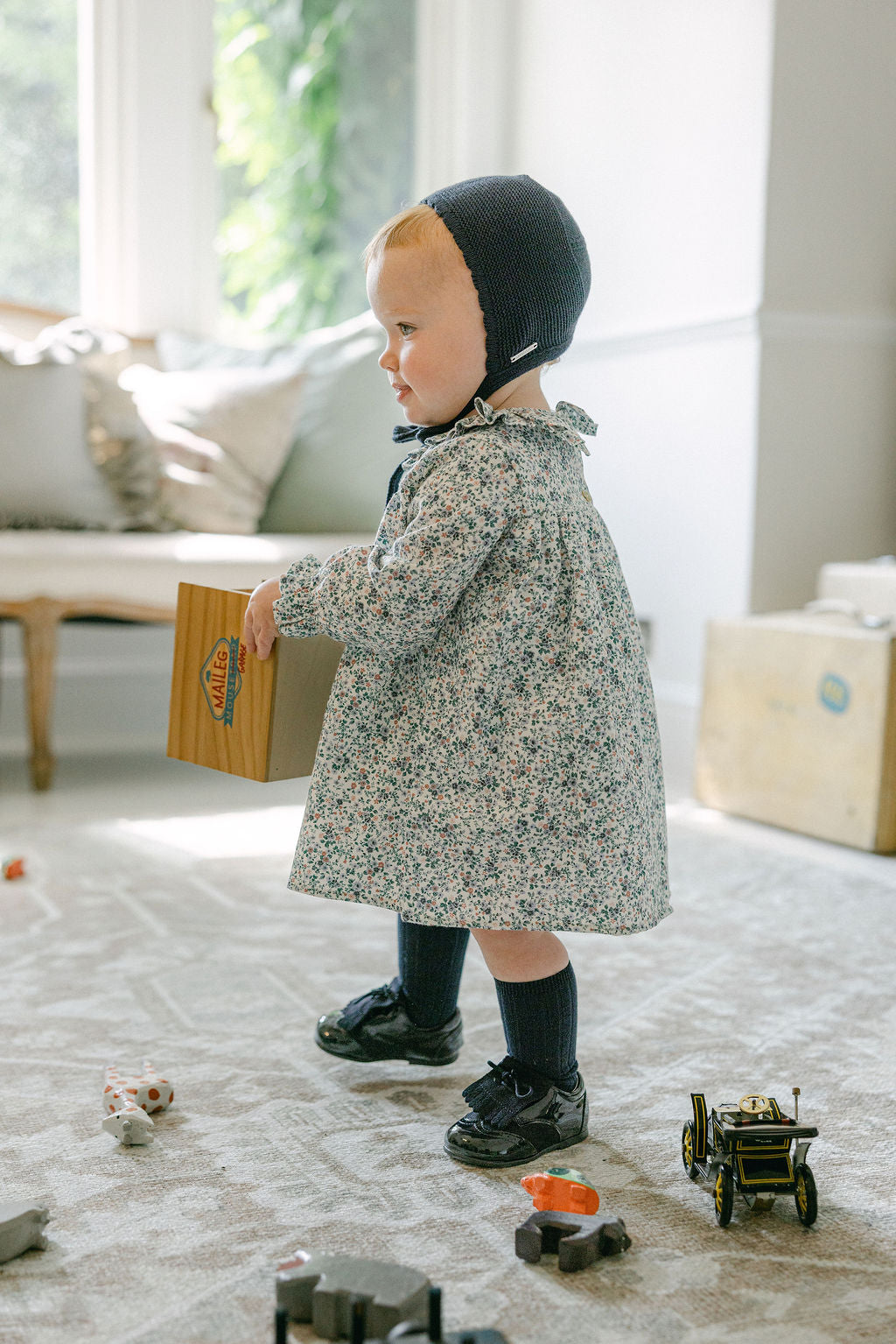 Child in a floral coat and black bonnet playing with toys on a carpeted floor.