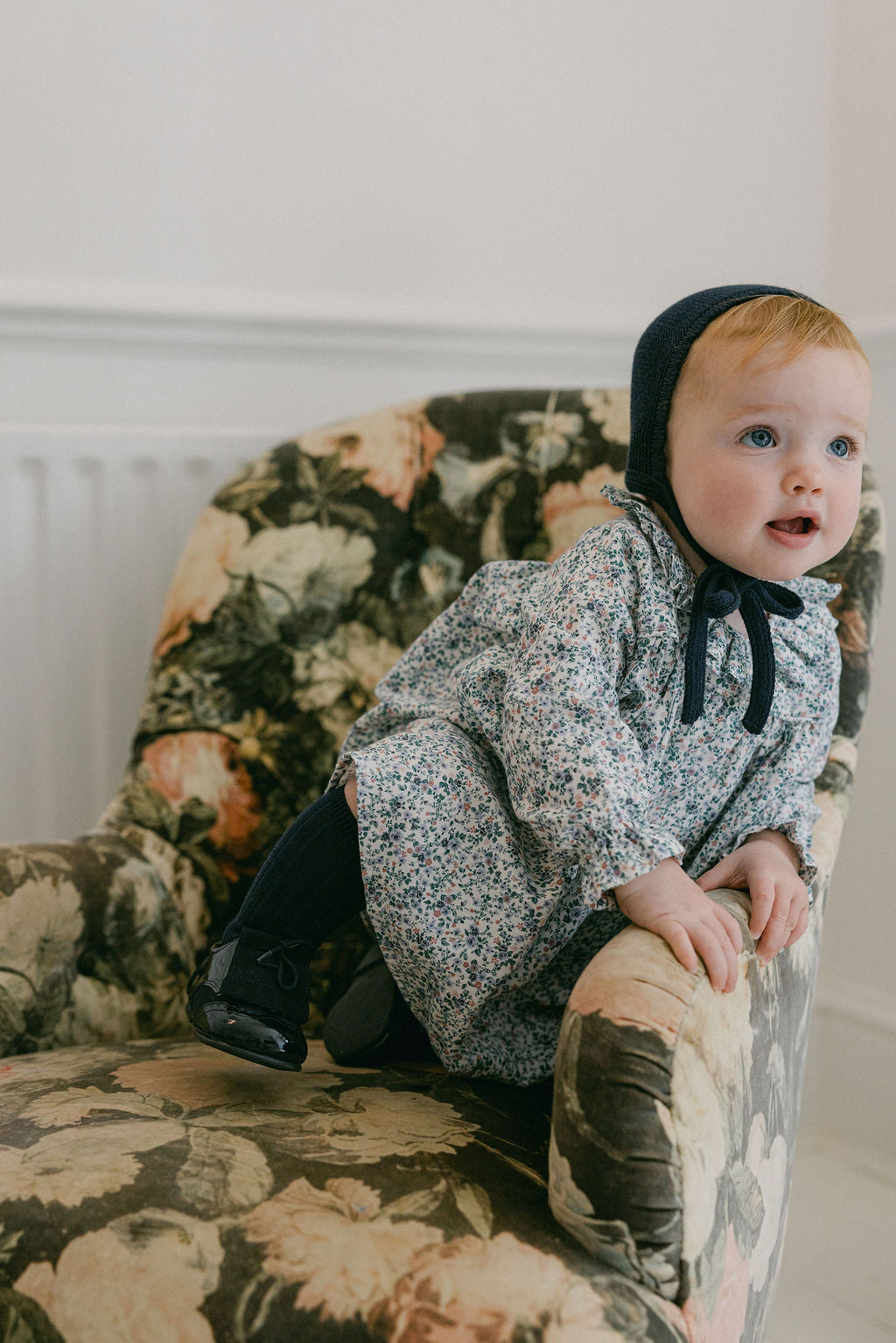 Baby in a floral armchair wearing a black bonnet