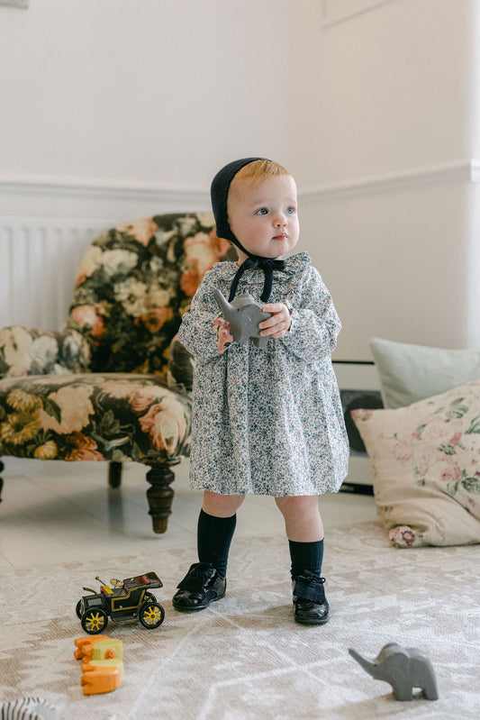 Child in a patterned dress standing in a room with floral armchair and toys on the floor.