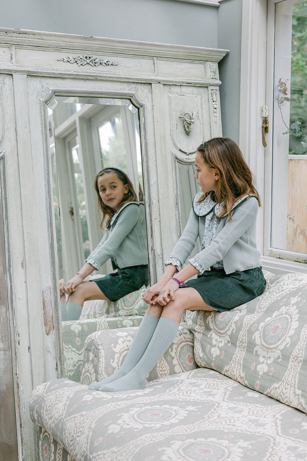 Young girl sitting on a patterned sofa in a room with a large mirror.