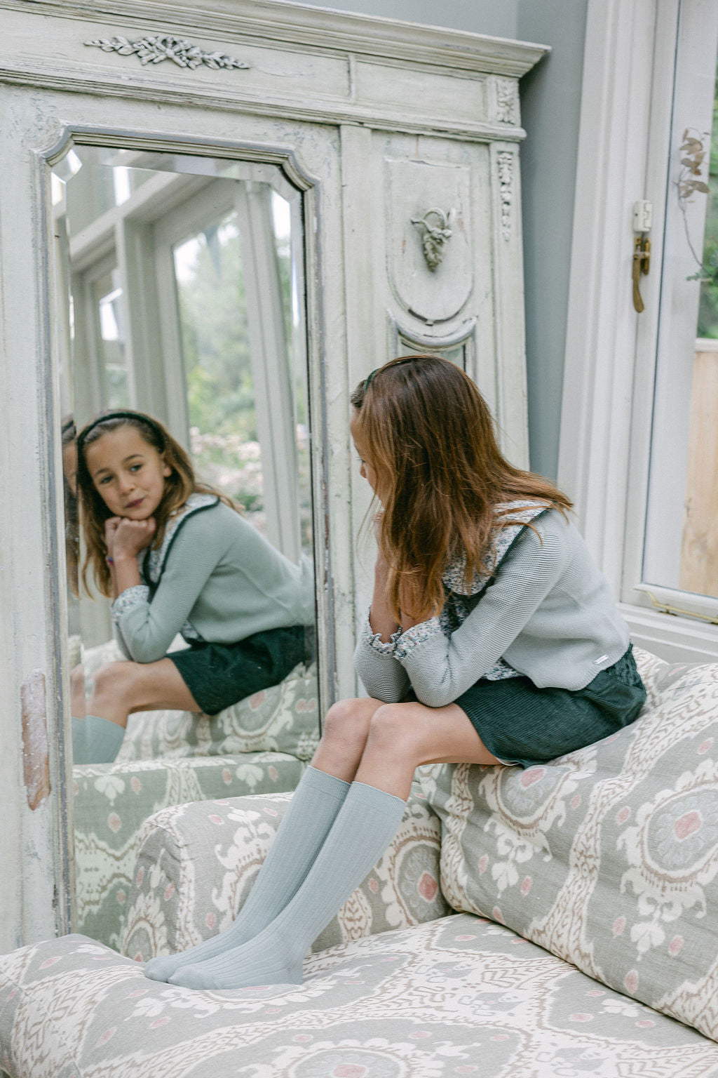 Young girl sitting on a bed wearing knee-high socks, looking at her reflection in a mirror.