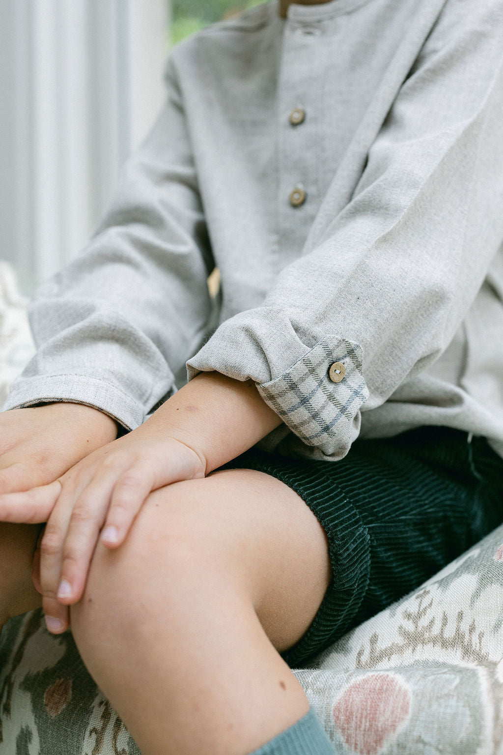 Person wearing a gray cardigan with rolled-up sleeves, sitting on a patterned surface.