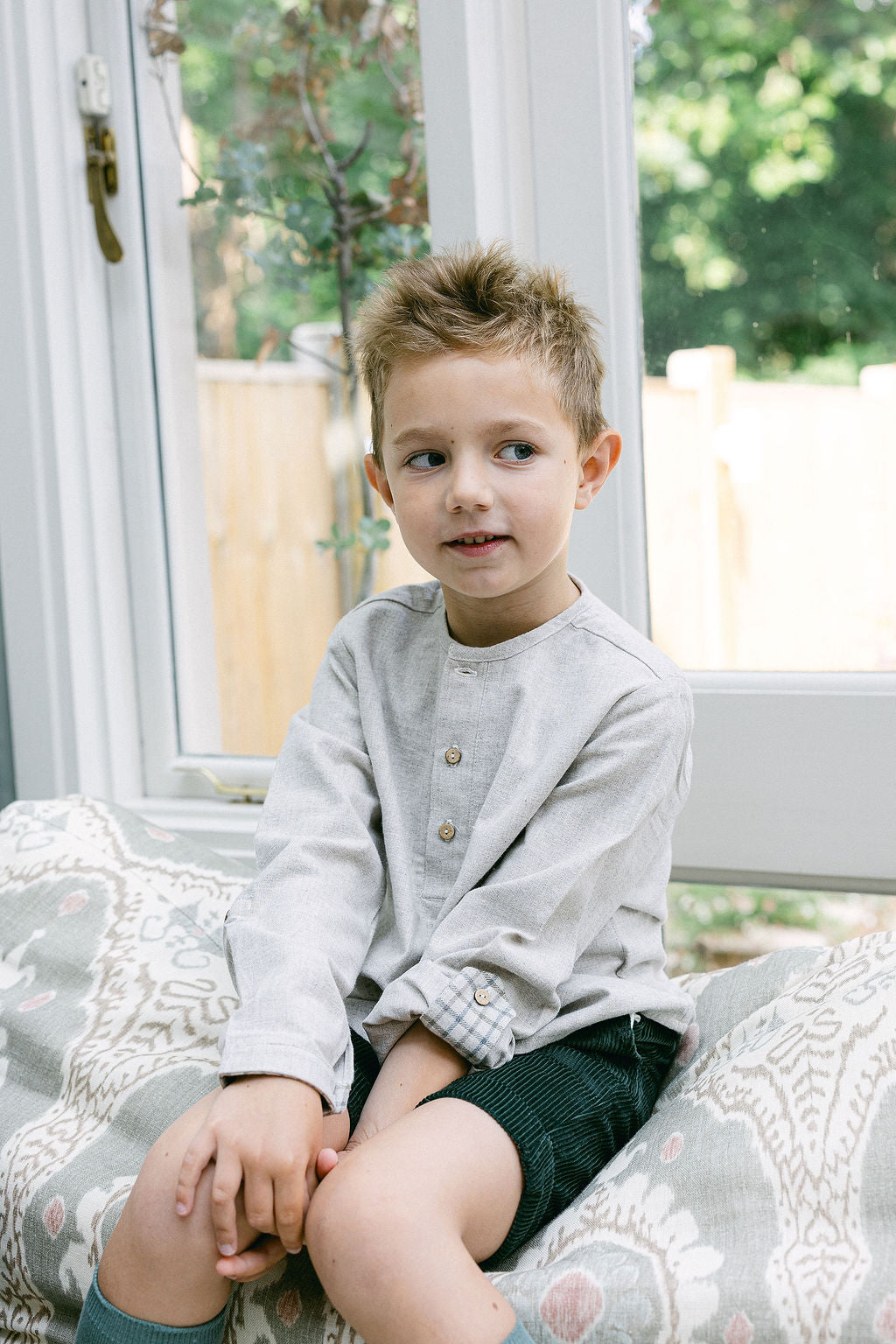 Young boy sitting on a patterned chair indoors with a window in the background