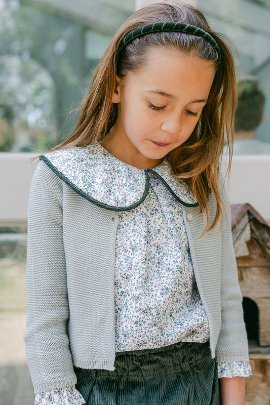Young girl wearing a light gray cardigan over a floral blouse with a blurred outdoor background