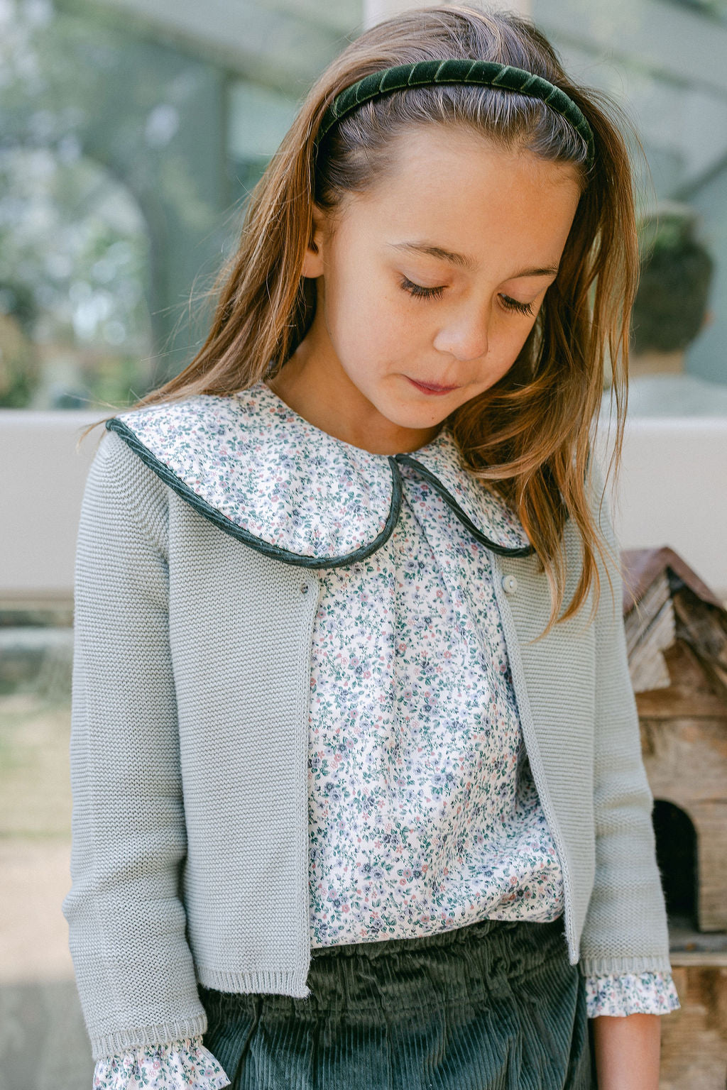 Young girl wearing a light gray cardigan over a floral blouse with a blurred outdoor background