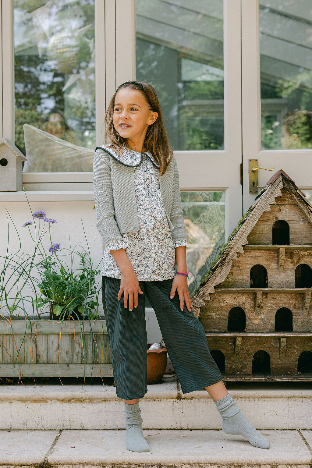 Young girl standing outdoors in front of a greenhouse, wearing a light gray jacket and dark green pants.