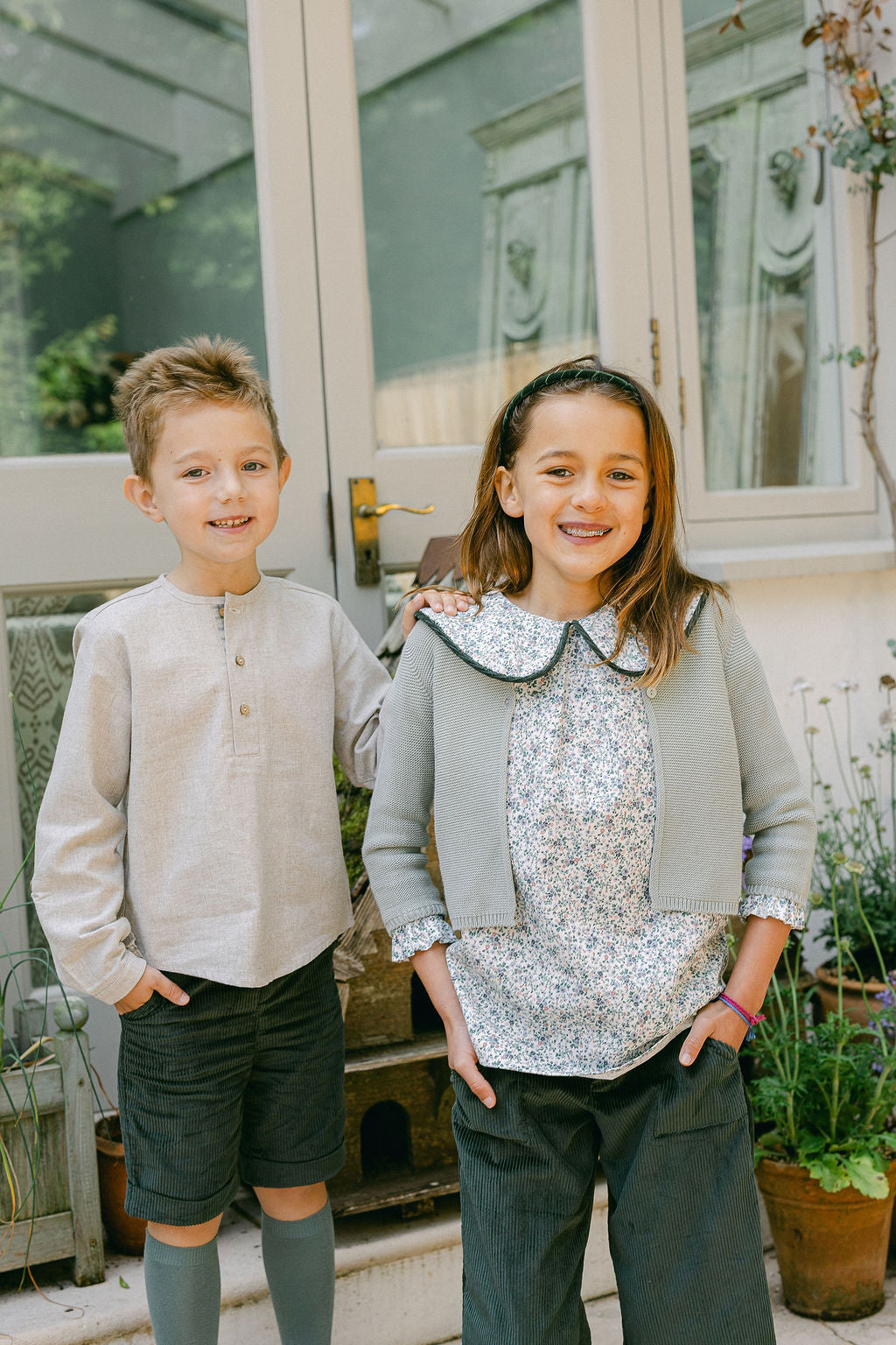 Two children standing outdoors in front of a house with plants around.