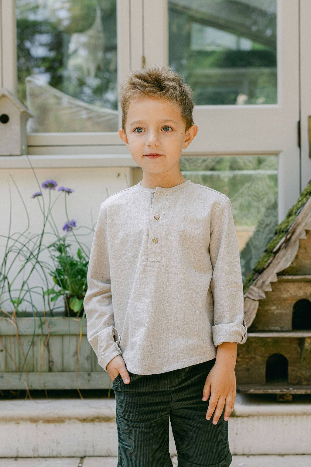 Young boy standing outdoors in front of a window with plants and a birdhouse.