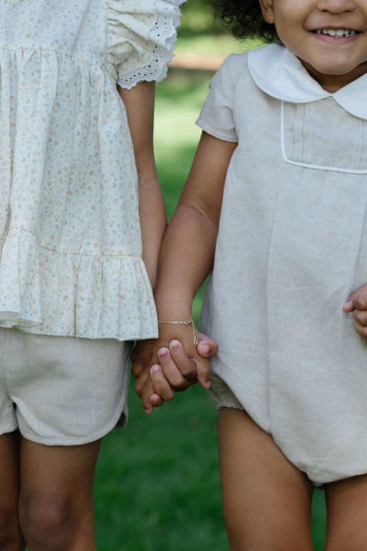 Two toddlers wearing beige linen rompers with Peter Pan collars, pleats on the front, and mother-of-pearl buttons.