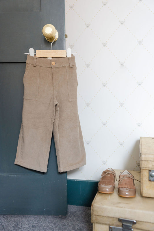 Brown pants hanging on a hook against a wall with shoes and a suitcase below.